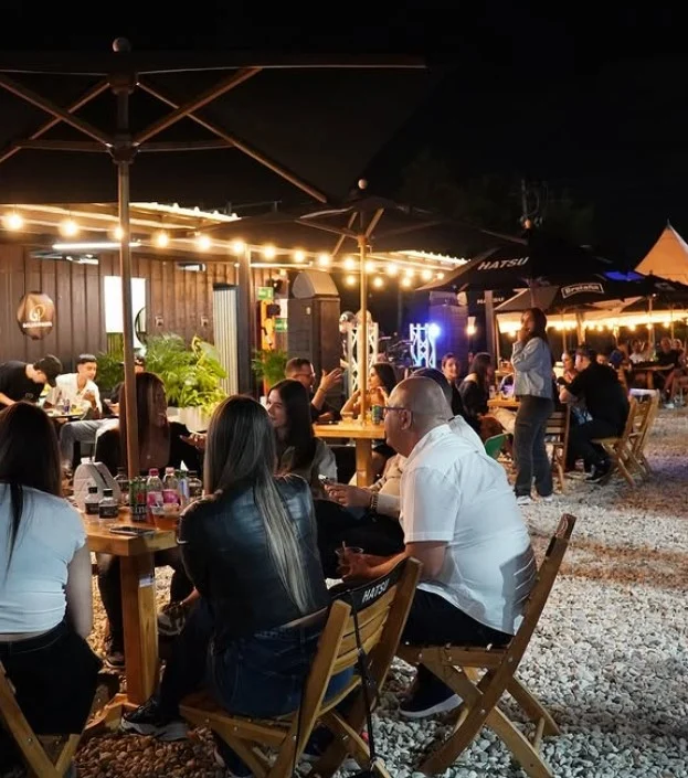 Espacio exterior con mesas de madera y sombrillas, iluminado con luces, en un ambiente social nocturno