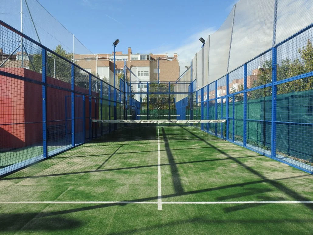 Cancha de pádel exterior con paredes de metal y césped artificial, rodeada de rejas y iluminación