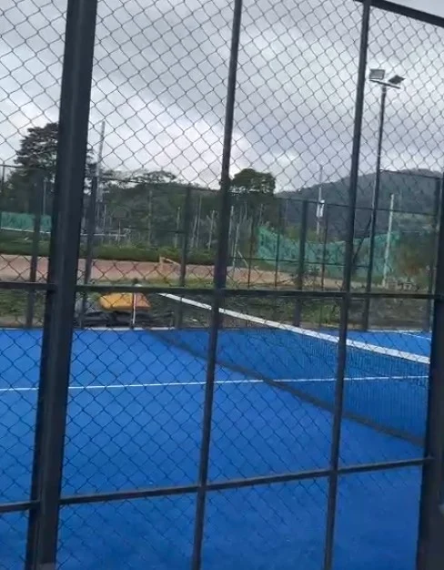 Rejas de metal en cancha de pádel exterior con superficie azul y fondo de canchas y árboles